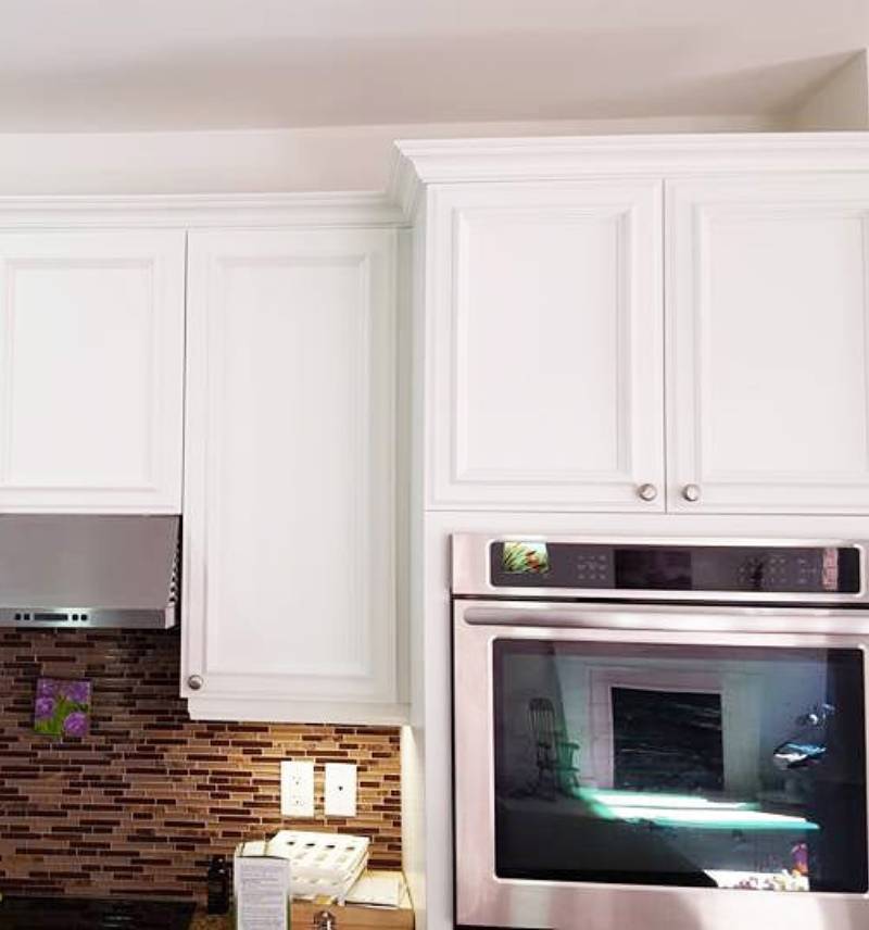 Freshly painted white kitchen cabinets with a smooth, high-quality finish, complemented by a modern stainless steel oven and a backsplash of brown and tan mosaic tiles, showcasing professional interior painting craftsmanship.